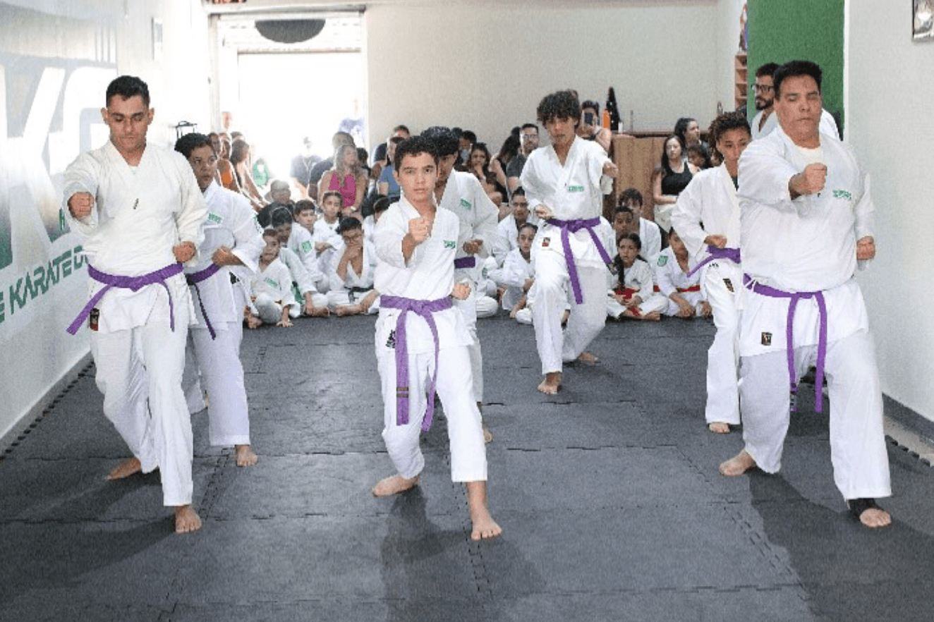 Treino de Kata na EKG: alunos de faixa roxa em posição de guarda executando um movimento sincronizado, com colegas e plateia ao fundo.